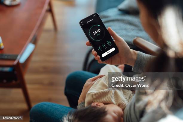 young woman using smart phone to adjust smart thermostat for air-conditioning at home while holding her baby to sleep - home appliances ストックフォトと画像