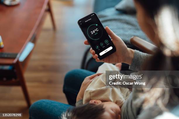 young woman using smart phone to adjust smart thermostat for air-conditioning at home while holding her baby to sleep - thermostaat stockfoto's en -beelden