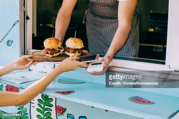 woman buying burgers from food van and using smartphone. - foodtruck stock pictures, royalty-free photos & images