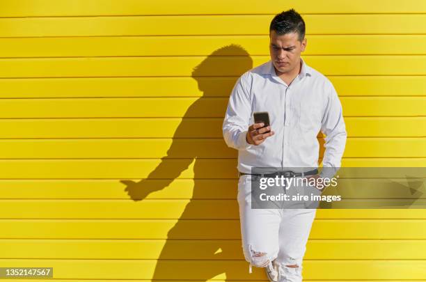 man standing leaning on yellow wall, checking his cell phone on a sunny day. - long sleeve stock pictures, royalty-free photos & images