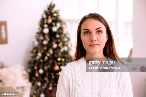 woman in white knitted sweater is looking at the camera against white interior with decorated christmas tree with garland. new year celebration concept. front view - knitted house stock pictures, royalty-free photos & images