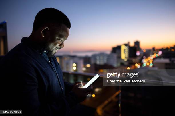 afrikanischer mann, der am frühen morgen auf dem balkon in der stadt e-mails am telefon überprüft - business man smartphone tablet stock-fotos und bilder