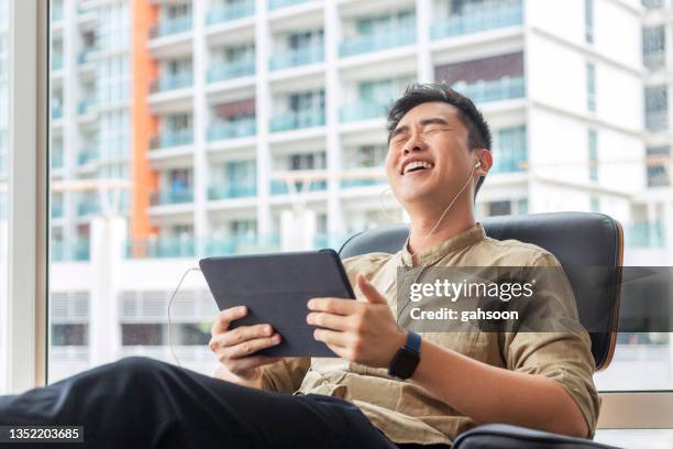 mann sitzt auf einer couch und lacht zu einem tablet im wohnzimmer - asian watching movie stock-fotos und bilder