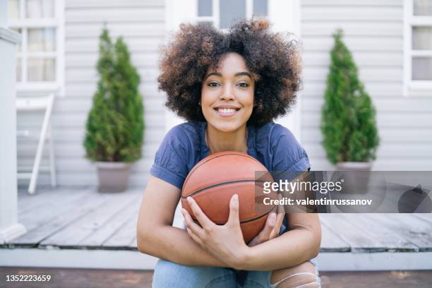 porträt einer lächelnden sportlerin mit afro-haaren. nahaufnahme einer jungen erwachsenen frau, die basketball hält und an einem sonnigen tag auf einer veranda sitzt. - female afro amerikanisch portrait stock-fotos und bilder