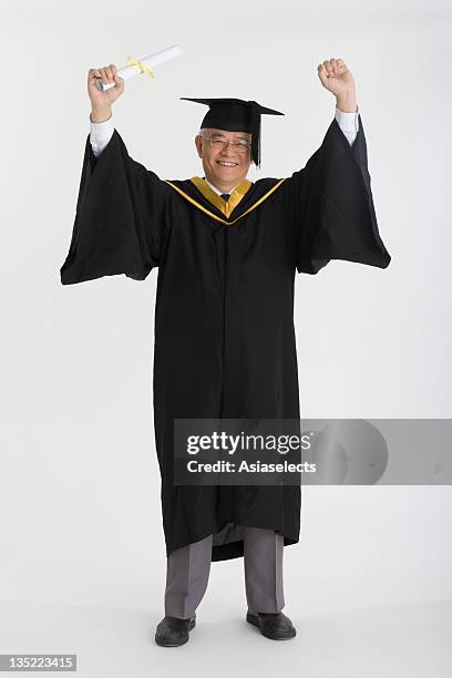 portrait of a senior man holding a diploma with his arms raised and smiling - paper gown stock pictures, royalty-free photos & images