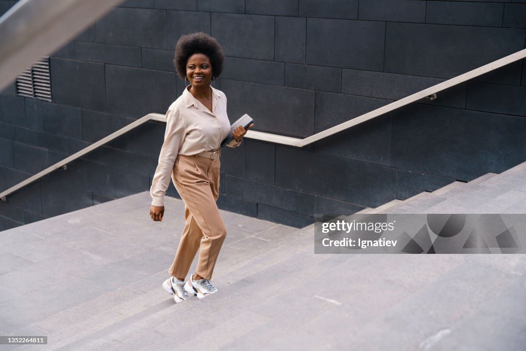 A smiling dark-skinned girl in a beige classical outfit with afro hairs, ascending the concrete stairs somewhere in a big city center