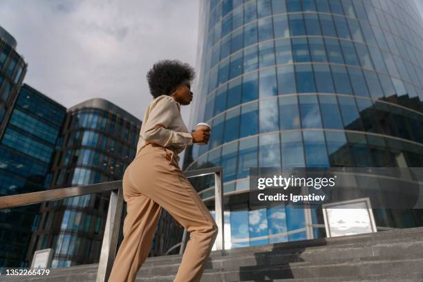 a low angle view on a dark skinned woman in beige outfit with african hairs ascending the stairs against the background with skyscrapers in a sunrise light - low confidence stock pictures, royalty-free photos & images