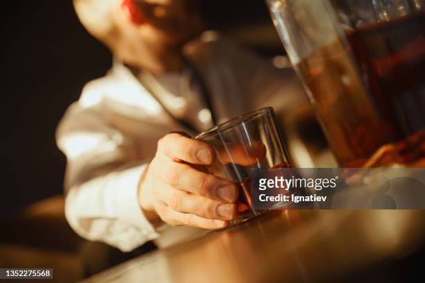 eine nahaufnahme der hand eines jungen mannes, dessen gesicht wir nicht sehen können, der ein glas whisky in einer dunklen bar mit der flasche im vordergrund hält - alkoholmissbrauch stock-fotos und bilder