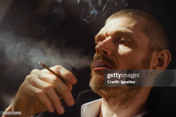 a close-up of a smoking man on a dark background in a warm light coming through the jalousie - premium lighter stock pictures, royalty-free photos & images