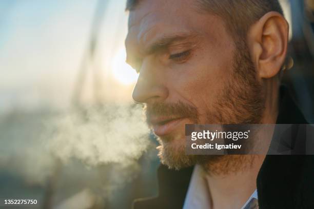close -up of a serious man smoking man looking down, standing on a roof during the dawn - stoppen met roken stockfoto's en -beelden
