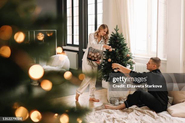 a young man and a woman in love decorate the christmas tree with balloons and toys on christmas day in the cozy living room of the home in winter. a couple in love in pajamas is preparing for the new year together in a country house - country christmas fotografías e imágenes de stock