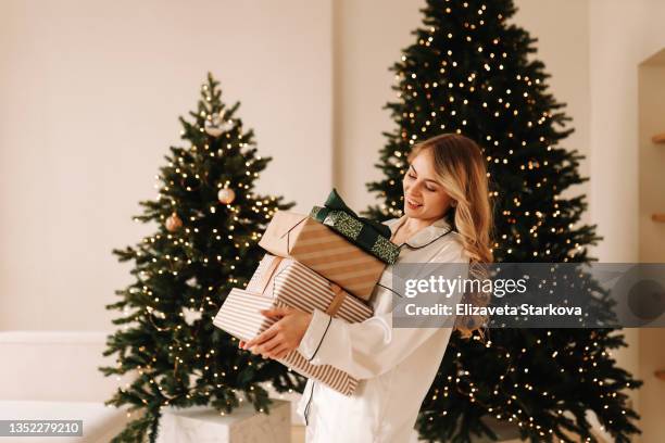 a happy smiling girl holds gift boxes and hides them under the christmas tree preparing for the new year holiday in a cozy decorated room of the indoor house - christmas present stockfoto's en -beelden