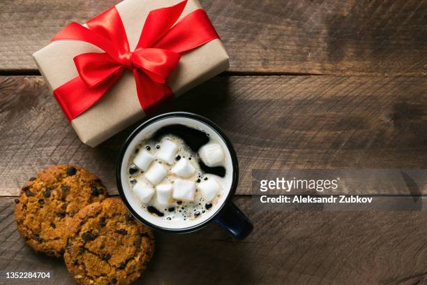the concept of preparing for a family holiday, happy christmas eve, valentine's day or birthday. wrapped in bright red ribbon christmas gift, on the background of a wooden table. a warming mug of hot coffee with marshmallow, cookie with chocolate. - hot chocolate band stock-fotos und bilder
