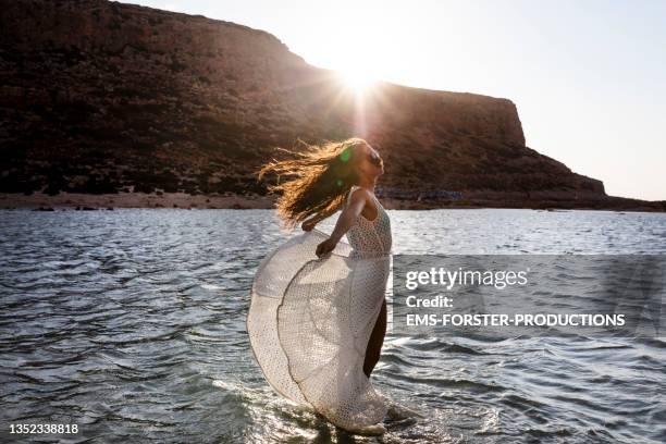 attractive woman with natural long brown curly hair wearing white sundress and sunglasses enjoys vacation time on the beach during sunset - white dress stock-fotos und bilder