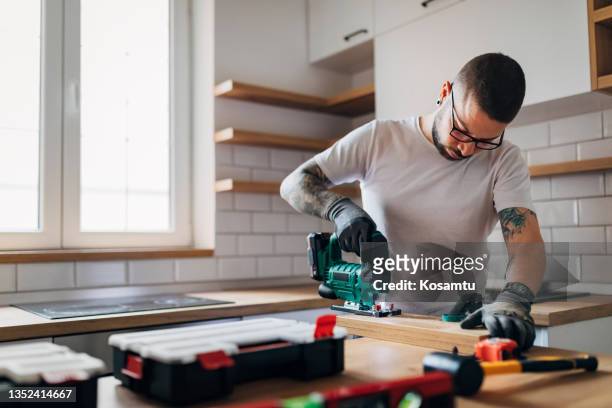 the bearded carpenter using electric jigsaw for cutting a wooden plank in his new kitchen. - elektrisch gereedschap stockfoto's en -beelden