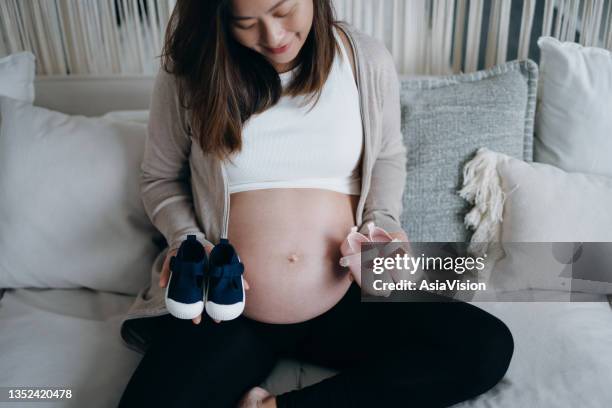 smiling young asian pregnant woman holding a pair of blue and pink baby shoes in front of her belly. expecting a new life, mother-to-be, gender reveal concept - chinese baby shoe stock pictures, royalty-free photos & images