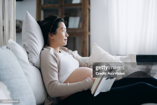 asiatische schwangere frau, die sich in einem gemütlichen zuhause entspannt und nachdenklich aussieht, während sie ein babybuch auf dem sofa liest. erwartung eines neuen lebens-, geburts- und erziehungskonzepts - pregnancy class stock-fotos und bilder