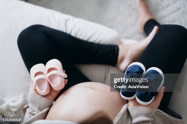 彼女の腹の前に青とピンクのベビーシューズを持っているアジアの妊婦の頭上のビュー。新しい人生を期待して、母親と、ジェンダーは概念を明らかにする - 赤ちゃんの靴 ストックフォトと画像