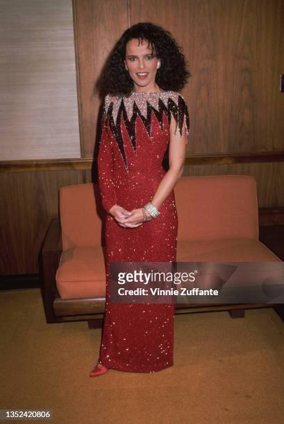 American actress and singer Shari Belafonte, wearing a red sequin asymmetric dress, stands before a small orange sofa, circa 1985.