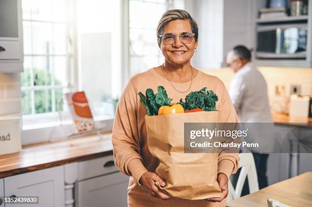 shot of a elderly woman holding a grocery bag in the kitchen - latin american and hispanic shopping bags stock pictures, royalty-free photos & images