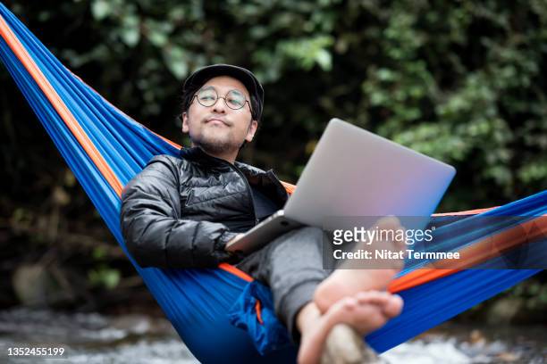 senior man lying in hammock using a laptop on a vacation. camping lover, nature addicted, and remote location. - hammock stock pictures, royalty-free photos & images