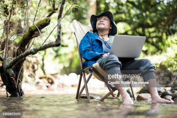 thoughtful senior man using a laptop while sitting on a camping chair at a stream in the forest. remote office, nature lover. - outdoor pursuit ストックフォトと画像
