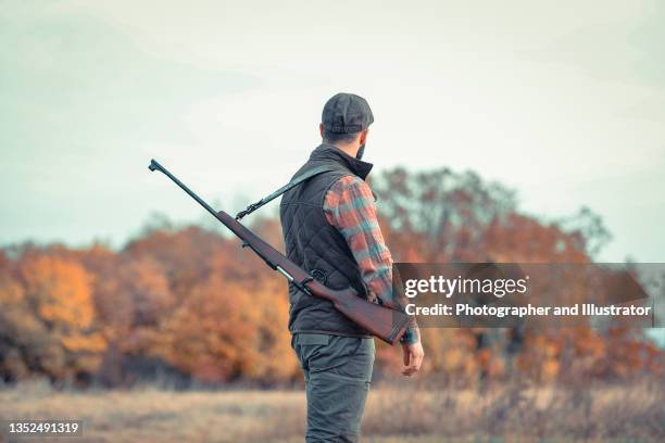 strong young hunter with black beard holding his gun and walking along the dirt road - hitman stock pictures, royalty-free photos & images