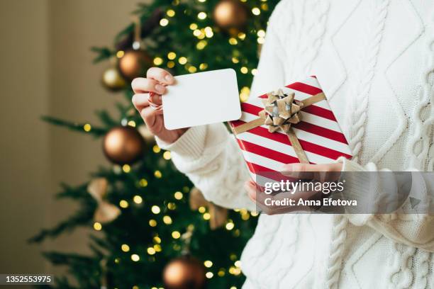 woman in white sweater is holding red-white gift box with gold bow and white tag against lights of decorated christmas tree. new year celebration concept. front view. close-up - gift tag and christmas stock pictures, royalty-free photos & images