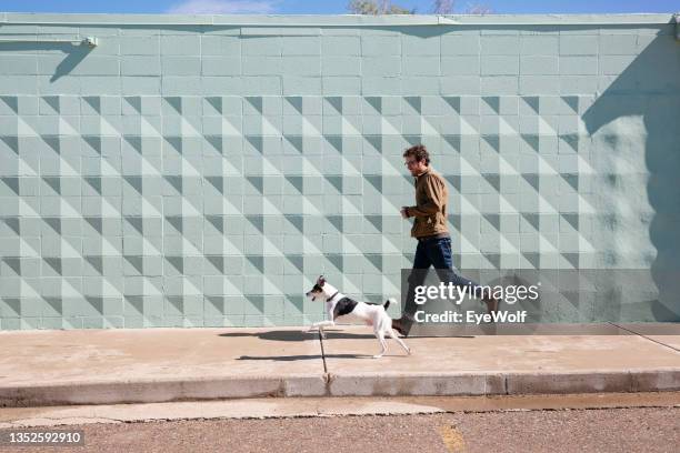 a man running with his dog on sidewalk in front of a blue texted wall. - city wall stock-fotos und bilder