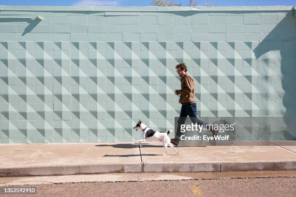 a man running with his dog on sidewalk in front of a blue texted wall. - vita cittadina foto e immagini stock