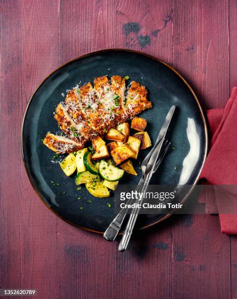 sliced schnitzel with roasted potatoes and zucchini on a plate on wooden red wooden background - schnitzel stockfoto's en -beelden