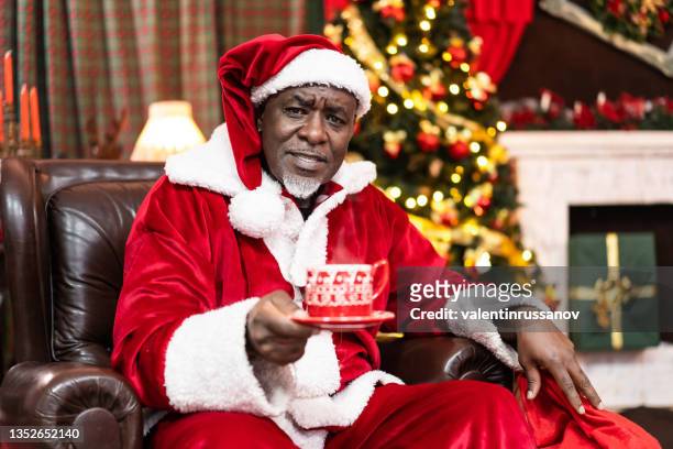 christmas and happy new year concept. kind looking african santa claus sitting on a chair near the christmas tree and holding a red cup, decorated with christmas ornaments, with hot drink  in it - happy new month stockfoto's en -beelden