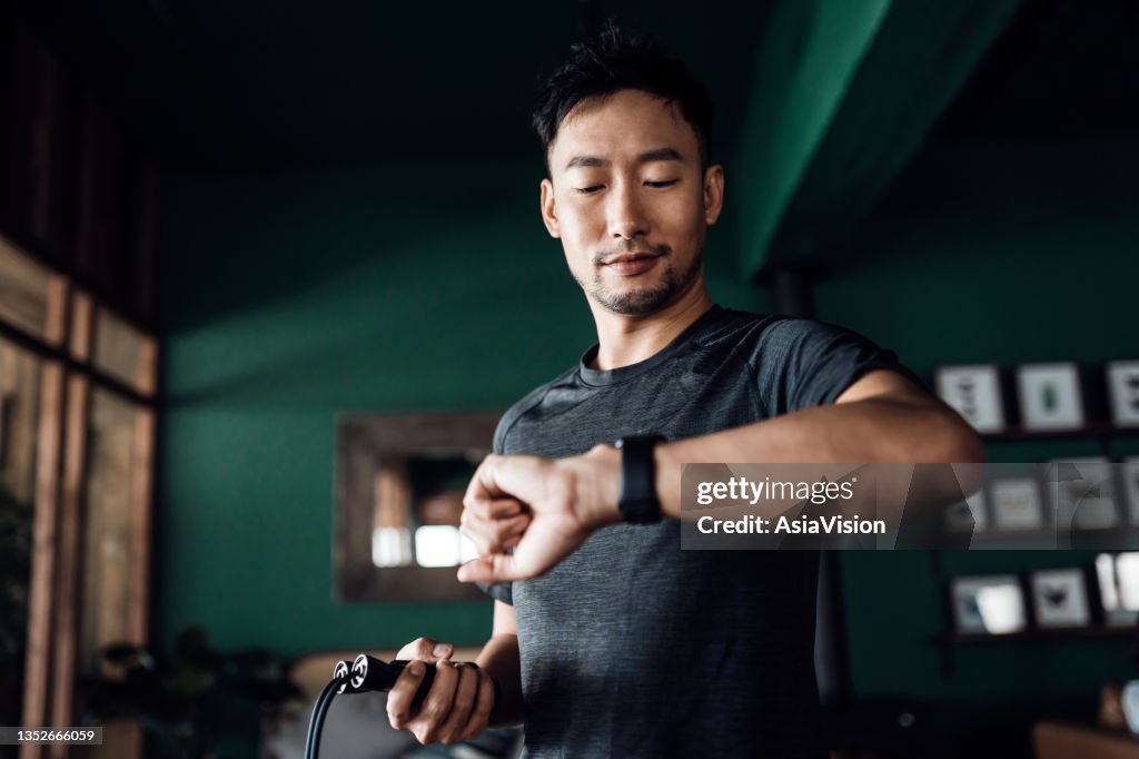 Active young Asian man exercising at home, using fitness tracker app on smartwatch to monitor training progress and measuring pulse. Keeping fit and staying healthy. Health, fitness and technology concept
