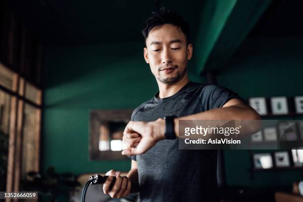 active young asian man exercising at home, using fitness tracker app on smartwatch to monitor training progress and measuring pulse. keeping fit and staying healthy. health, fitness and technology concept - indoor athletics stock pictures, royalty-free photos & images