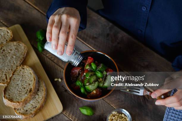 vegetarian, vegan, and raw food. a girl or woman eats a vegetable salad of tomatoes, cucumbers, nuts and seeds, green basil leaves, and salts it from a salt shaker. next to it are sliced baguette slices. the concept of healthy eating, diet. - gesalzenes stock-fotos und bilder