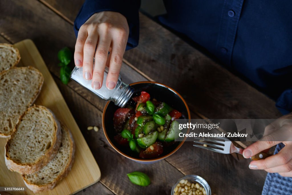 Vegetarian, vegan, and raw food. A girl or Woman Eats a Vegetable salad of tomatoes, cucumbers, nuts and seeds, Green Basil Leaves, and Salts It From a salt Shaker. Next To It Are Sliced Baguette Slices. The concept of Healthy eating, diet.