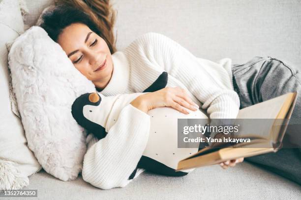 beautiful sleepy woman is reading a book on a cozy sofa hugging her plush penguin toy - comfort zone ストックフォトと画像