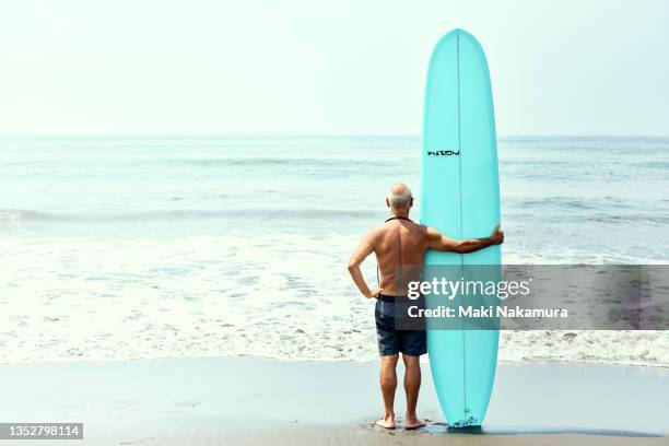 a portrait of a senior man in the back standing on the beach with a surfboard. - pension concept stock pictures, royalty-free photos & images