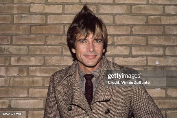 American actor Dirk Benedict wearing a tweed jacket over a checked shirt and tie, posing against a bare brick wall, circa 1985.