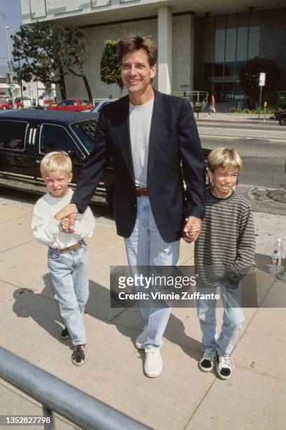 American actor Dirk Benedict, wearing a dark blue blazer over a white t-shirt with stonewashed jeans, holding hands with his two sons, George and...