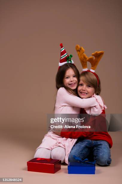 portrait of beautiful cute boy sitting celebrating christmas dressing red knitted jumper and blue jeans and christmas red socks bezel with deer antlers and beautiful girl dressing pink dress and white tights   on beige background - red dress child stockfoto's en -beelden