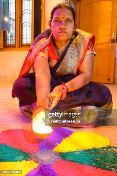 image of indian woman placing lit diya (oil lamp) at centre of gulal rangoli design, red, purple, yellow and green powder paint flower petal pattern, flickering flame, diwali festival of lights celebration - diya oil lamp stockfoto's en -beelden