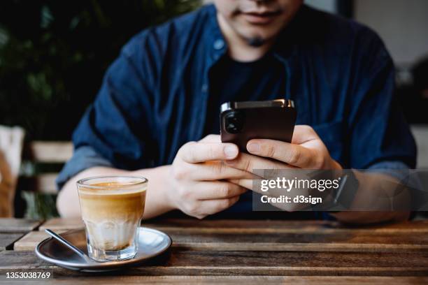cropped shot, mid-section of young asian man sitting at a table in cafe, using smartphone while having a cup of coffee. lifestyle and technology - paypal stock pictures, royalty-free photos & images