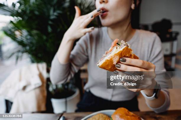 cropped shot, mid-section of young asian woman eating freshly made delicious cheeseburger in a cafe, licking her fingers. enjoying her lunch! people and food concept - delicious ストックフォトと画像