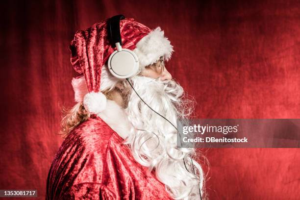 person dressed as santa claus, in profile, with white headphones listening to music, on red vintage background. christmas, santa claus, gifts, december and celebrations concept. - weihnachtsmusik stock-fotos und bilder