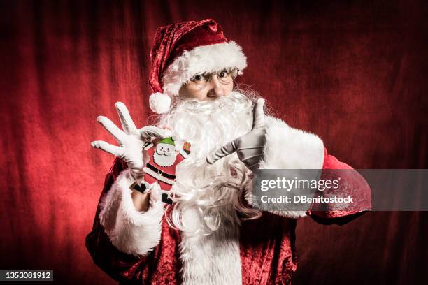 person dressed as sana claus, pointing to a small christmas ornament, on red vintage background. christmas, santa claus, gifts, december and celebrations concept. - naughty santa 個照片及圖片檔