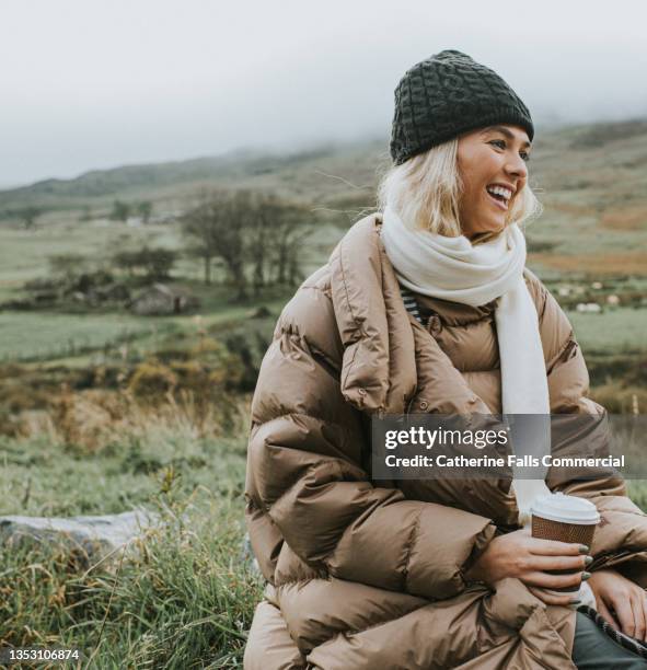 a beautiful young woman clutches a takeaway coffee cup, as she enjoys being outdoors - padded jacket 個照片及圖片檔