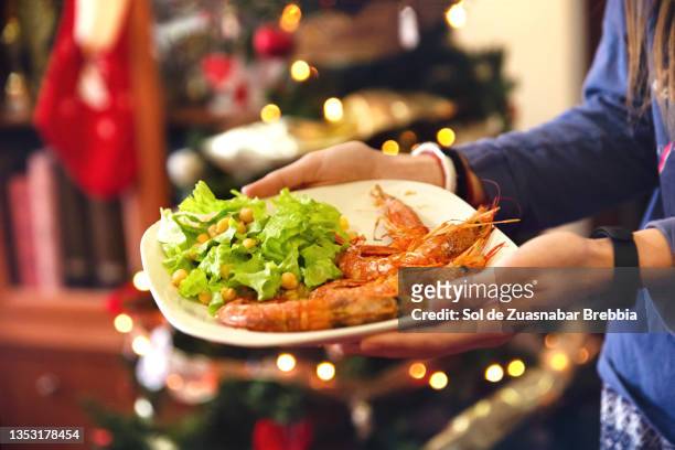 close-up of a plate with salad and prawns held in the hand of a teenage girl who brings it to the christmas table. behind you see the bokeh of the christmas tree lights - gamba marisco - fotografias e filmes do acervo