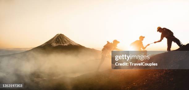 group of hikers on a mountain. man helping her friend to climb a rock. young asian three hikers climbing up on the peak of mountain near mountain fuji. giving a helping hand. climbing. helps and team work concept - people climbing walking mountain group stock pictures, royalty-free photos & images