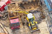 Excavator digs a hole and loads soil into the back of a truck, aerial top view.